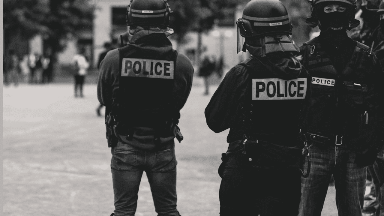 Policajti v taktickom vybavení na námestí, čiernobiela fotografia mestského prostredia so zameraním na bezpečnosť.