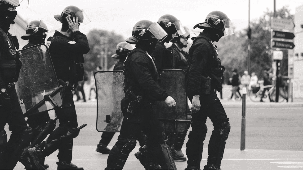 Policajti v zásahovej výstroji so štítmi kráčajú po ulici počas verejnej demonstrácie.