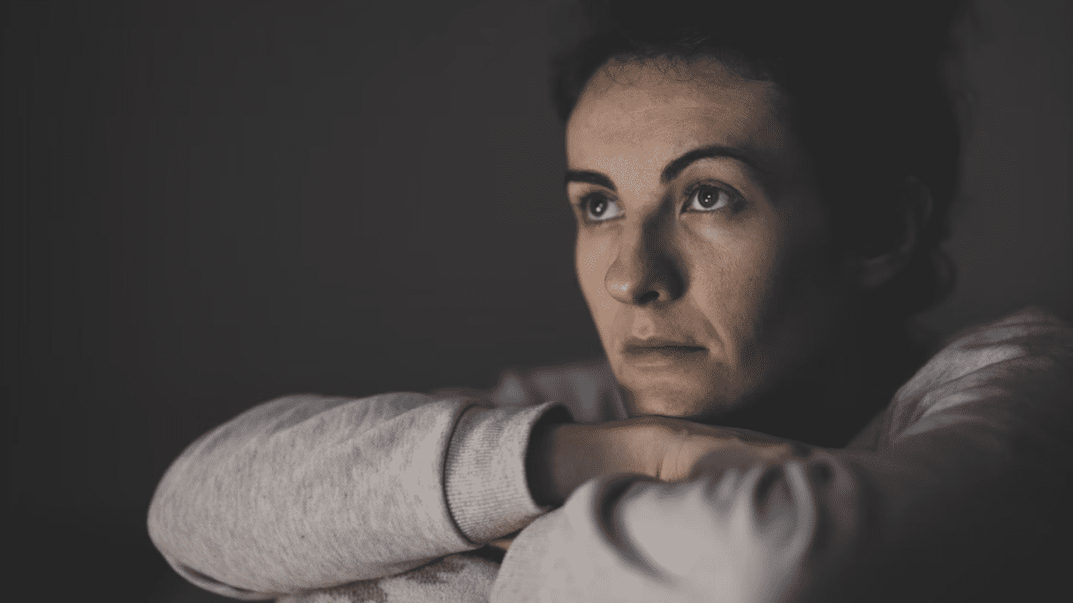 Žena v šedom svetri zamyslene pozerá do diaľky na tmavom pozadí.