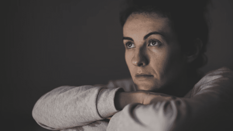 Žena v šedom svetri zamyslene pozerá do diaľky na tmavom pozadí.