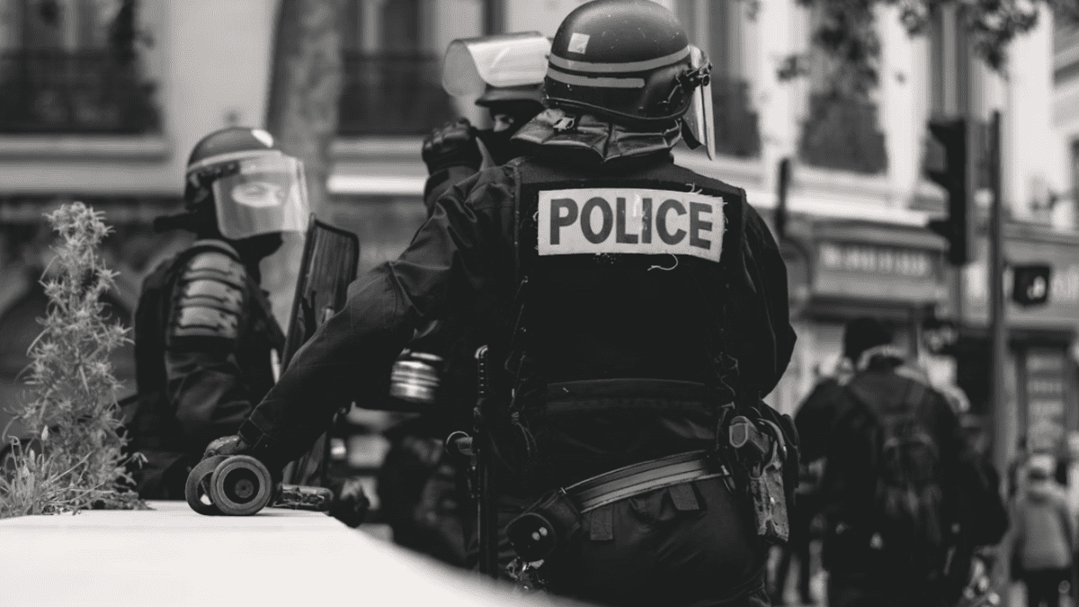 Policajti v ochranných uniformách na hliadke počas mestského zásahu, čiernobiely pohľad zozadu.