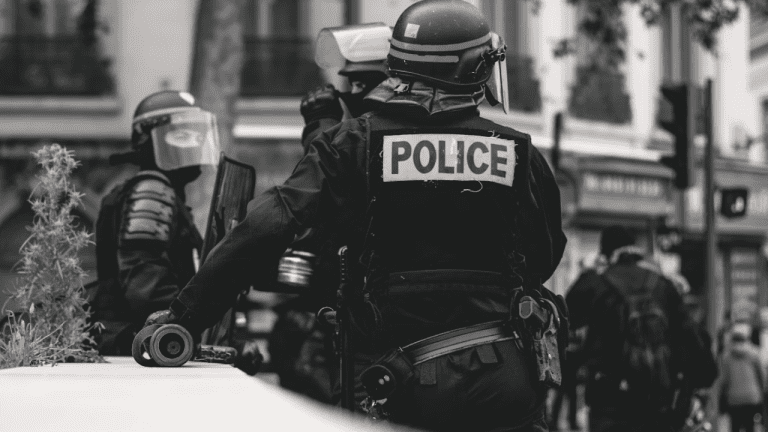 Policajti v ochranných uniformách na hliadke počas mestského zásahu, čiernobiely pohľad zozadu.