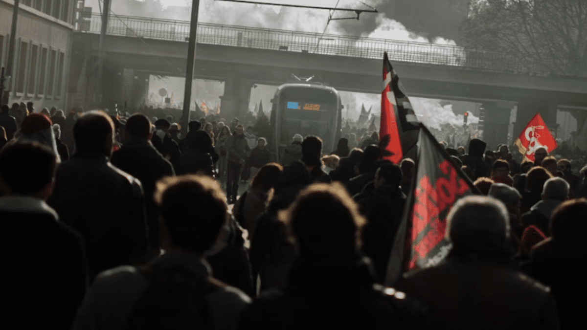 Dav ľudí pochoduje ulicou s vlajkami, v pozadí tramvaj a most. Hustá atmosféra demonštrácie.