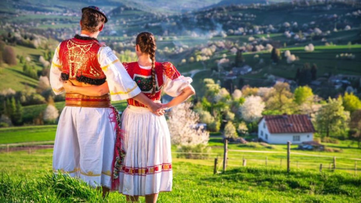 Pár v tradičných slovenských krojoch obdivuje malebnú krajinu s rozkvitnutými stromami a domom v údolí.