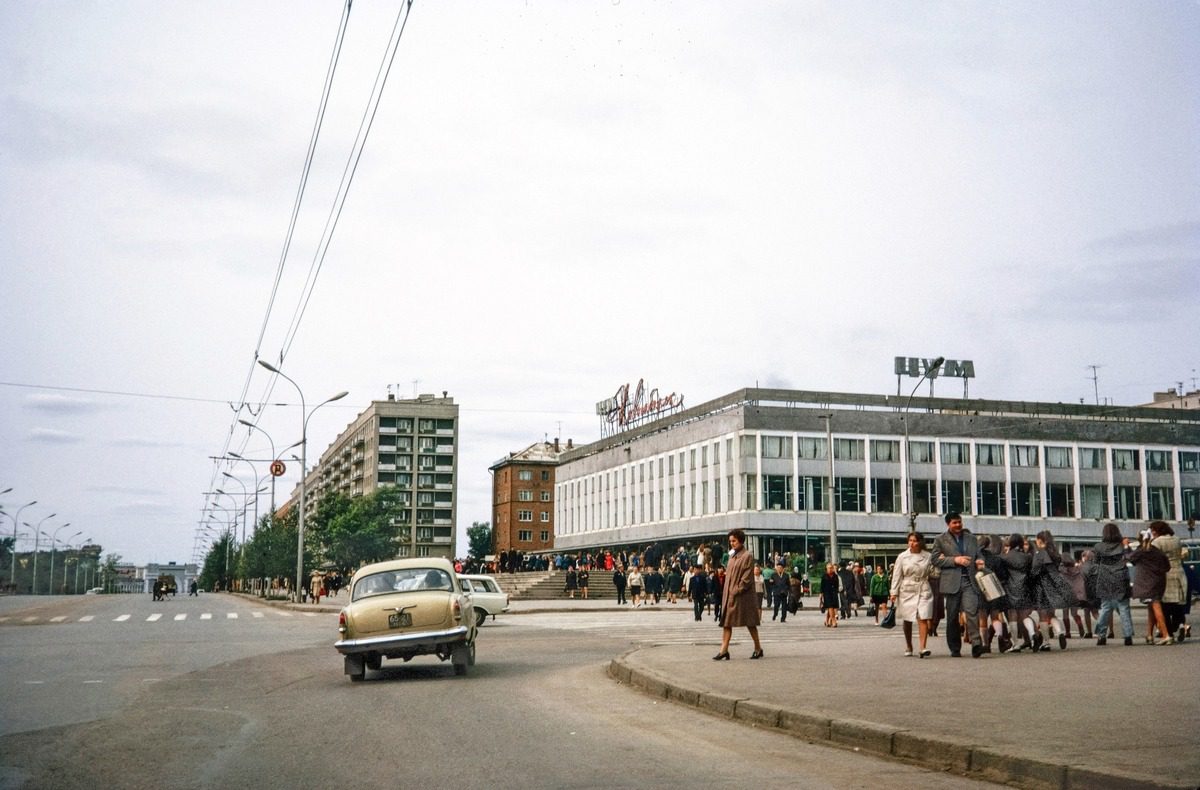 Historická ulica s automobilom a chodcami pred veľkou budovou. Retro scenéria mesta za jasného dňa.