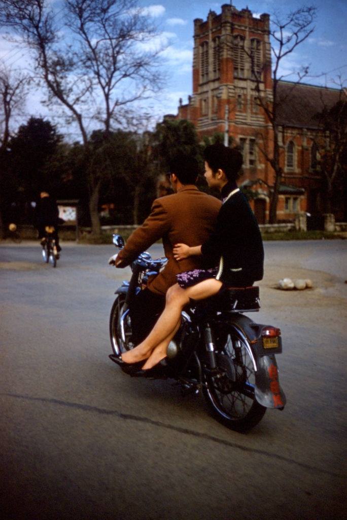 Pár jazdí na motocykli v blízkosti historickej budovy so stromami v pozadí.