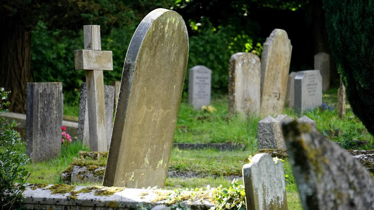 Starý cintorín s machom pokrytými náhrobnými kameňmi a dreveným krížom v zelenom prostredí.