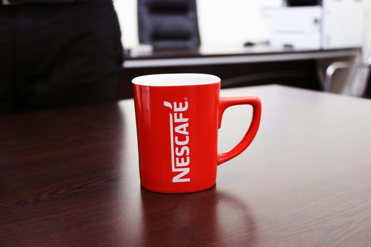 Červený hrnček s logom Nescafé na drevenom stole v kancelárii, pripravený na chvíle pohody s kávou.