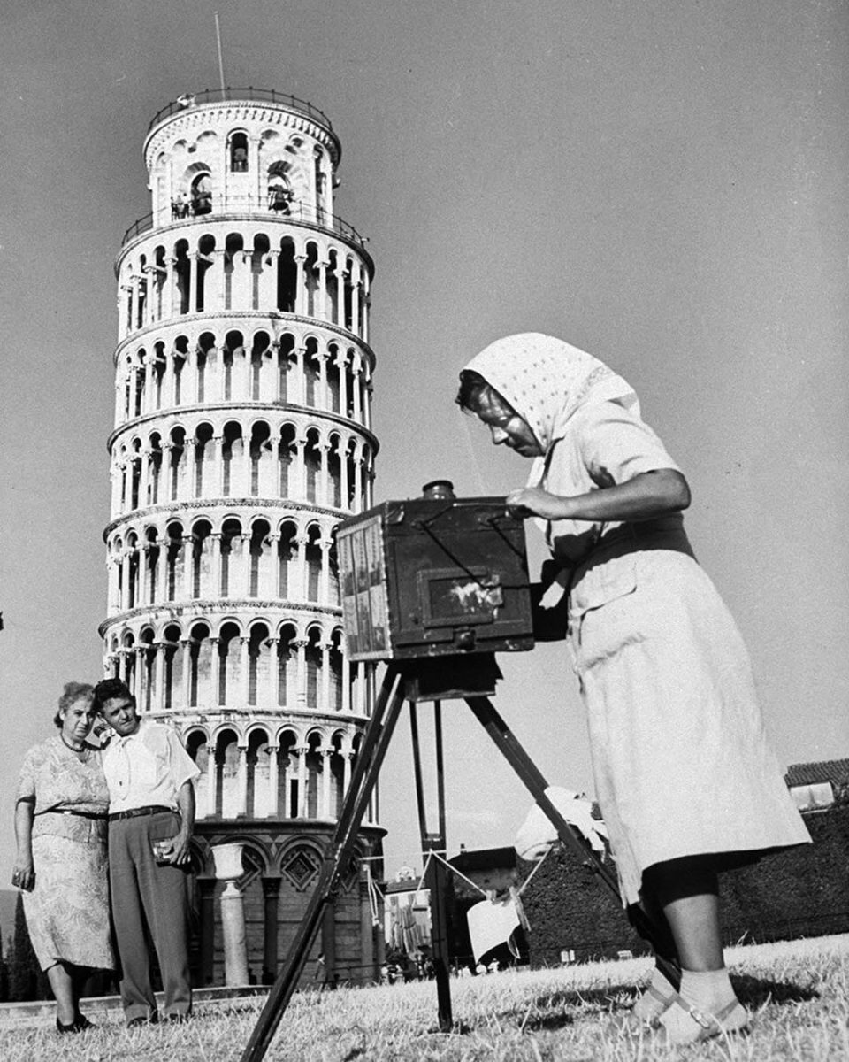 Fotografka s veľkým fotoaparátom fotí pár pred šikmou vežou v Pise, čiernobiely historický moment.