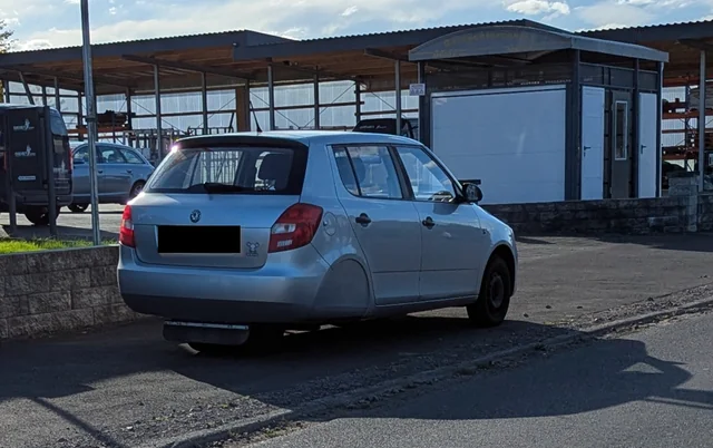 Strieborné auto zaparkované pri ceste pred skladovou budovou v jasnom, slnečnom dni.