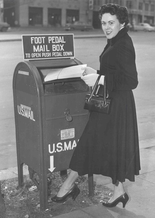Žena v kabáte vkladá list do historickej americkej poštovej schránky s pedálom na rušnej ulici.