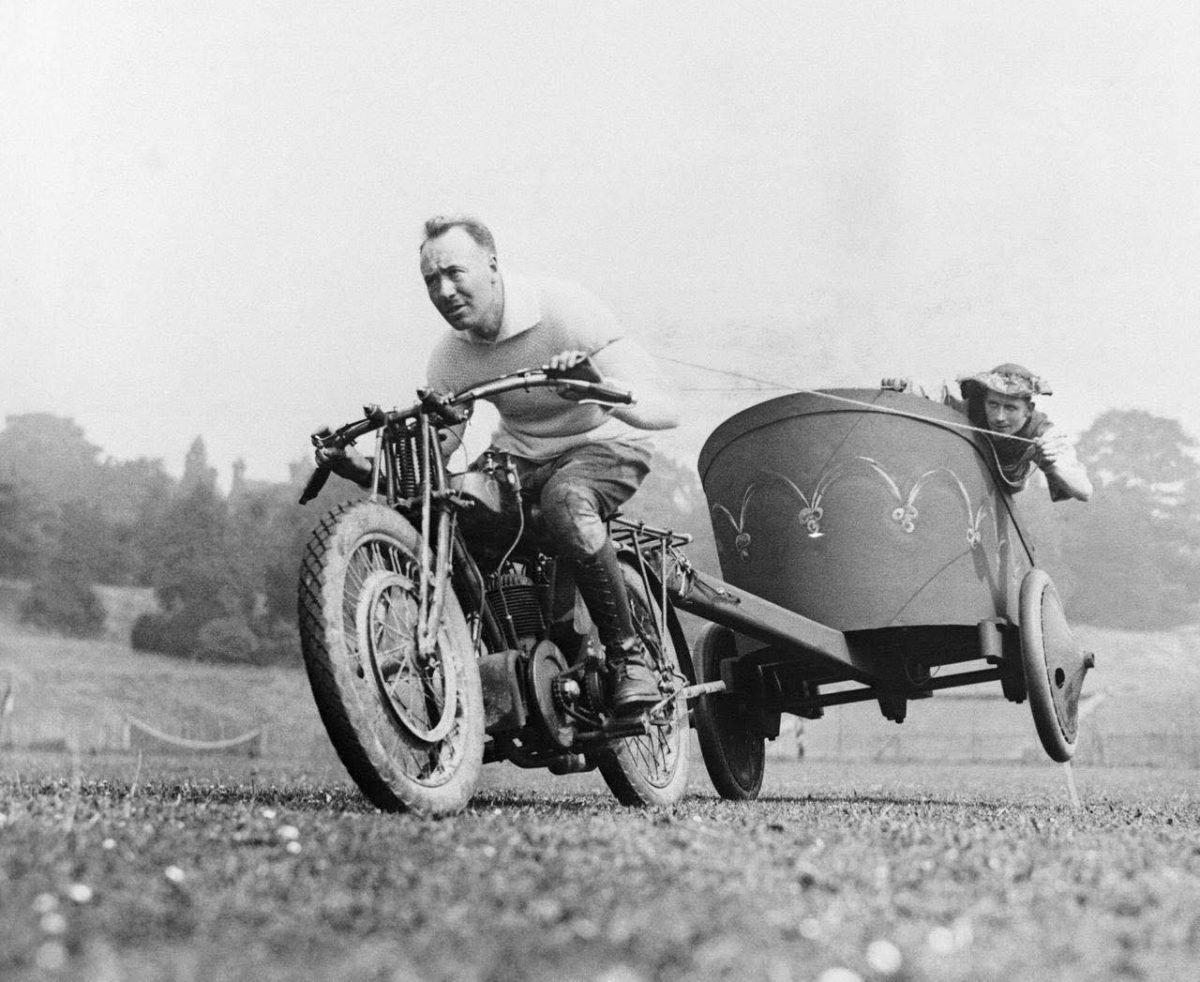 Muž na motorke ťahá vozík s človekom po lúke. Vintage scéna rýchlej jazdy v prírode.