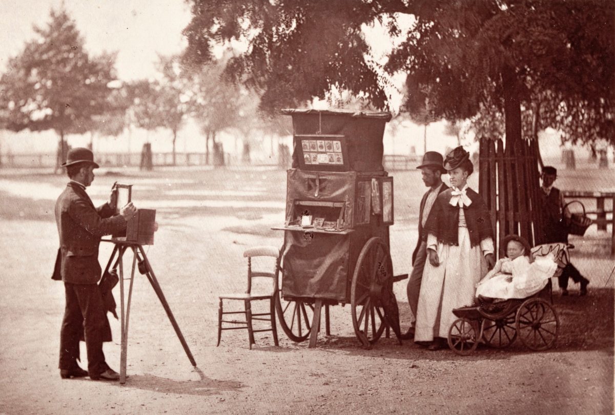 Historická fotografia z parku: fotograf s kamerou na statíve a rodina s kočíkom pózuje pod stromami.