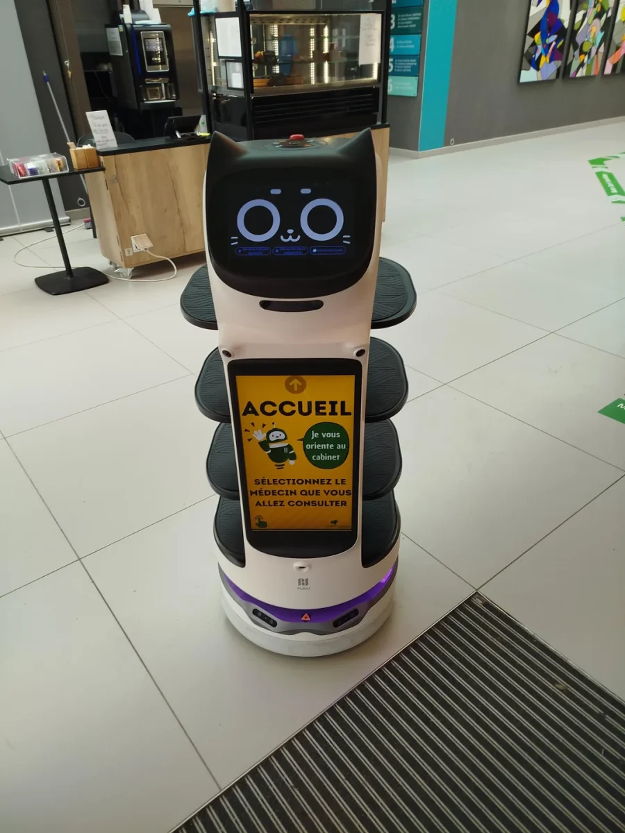 Robot s obrazovkou v tvare mačky v modernom priestore so smerovým nápisom pre návštevníkov.
