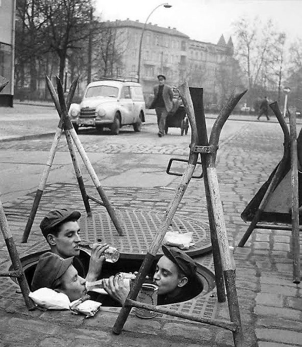 Ľudia oddychujú v poklope na ulici, popíjajú nápoje. V pozadí staré auto a budovy. Komická scénka z minulosti.
