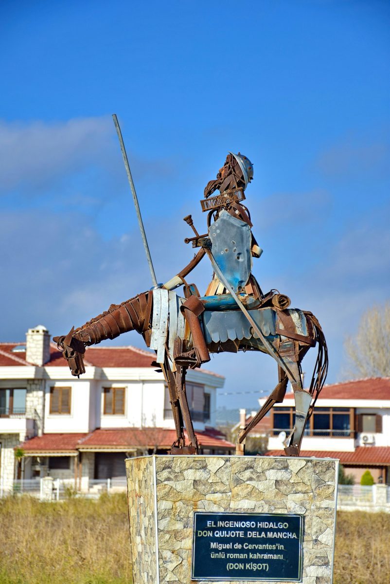 Socha Dona Quijota z kovového odpadu na koni, modrá obloha v pozadí, dedinská zástavba naokolo.