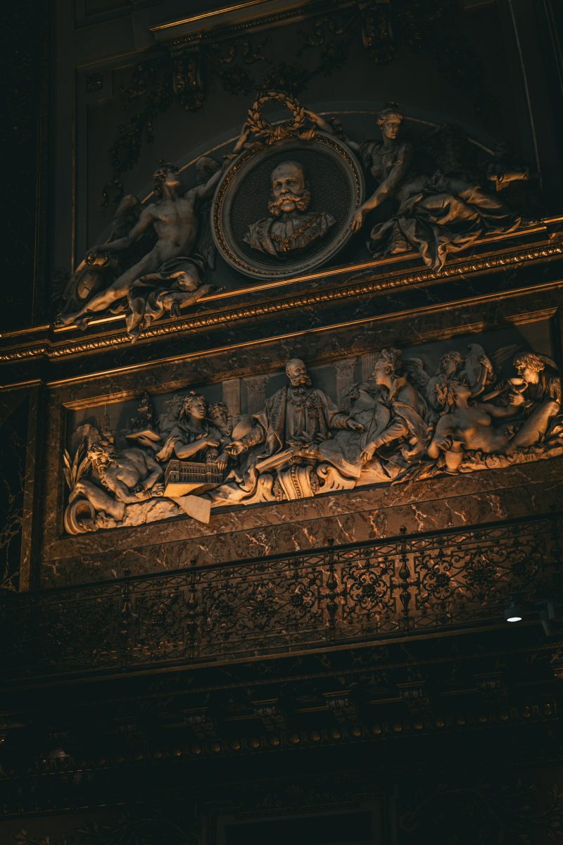 Historický reliéf na stene s postavami a okrúhlym portrétom osvetlený teplými tónmi svetla.