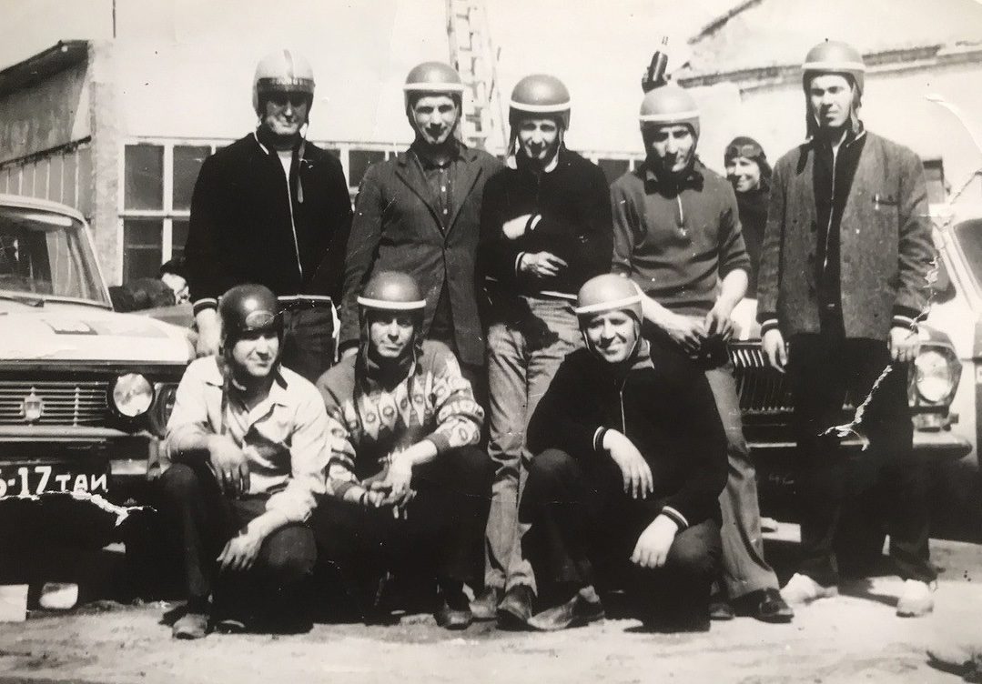 Skupina mužov v prilbách pred autami na historickej fotografii z 20. storočia. Retro motoklub.