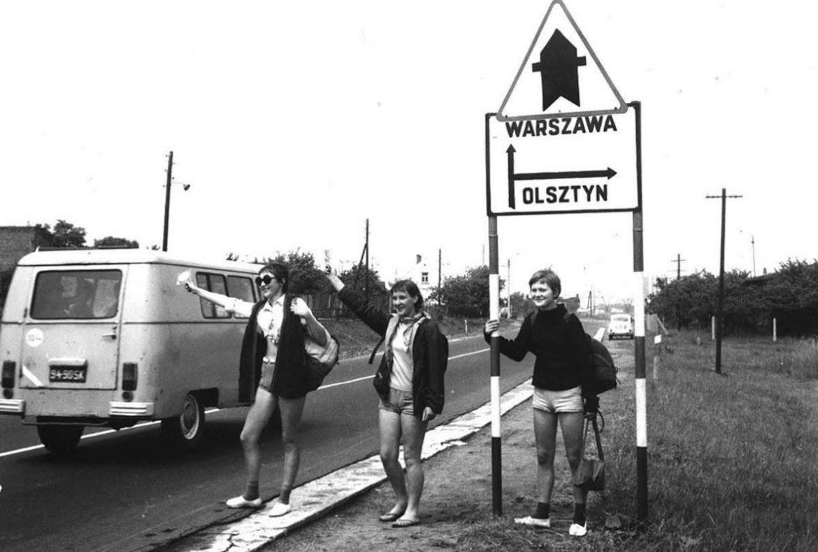 Traja stopári na ceste pod značkou, smer Varšava a Olsztyn, s prechádzajúcou dodávkou na vidieckej ceste.