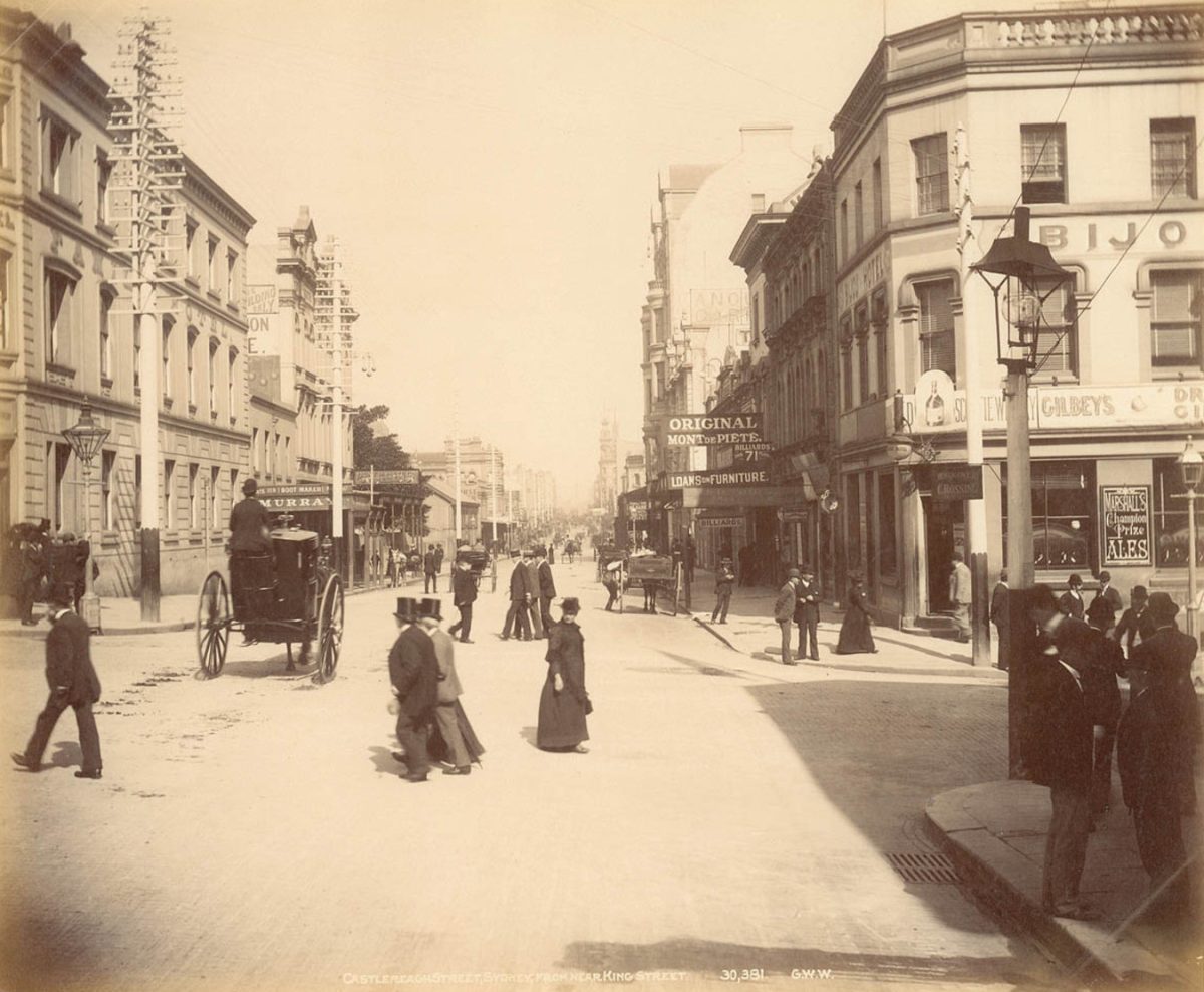 Historická fotografia rušnej ulice s kočmi a chodcami, obklopená budovami a obchodmi.