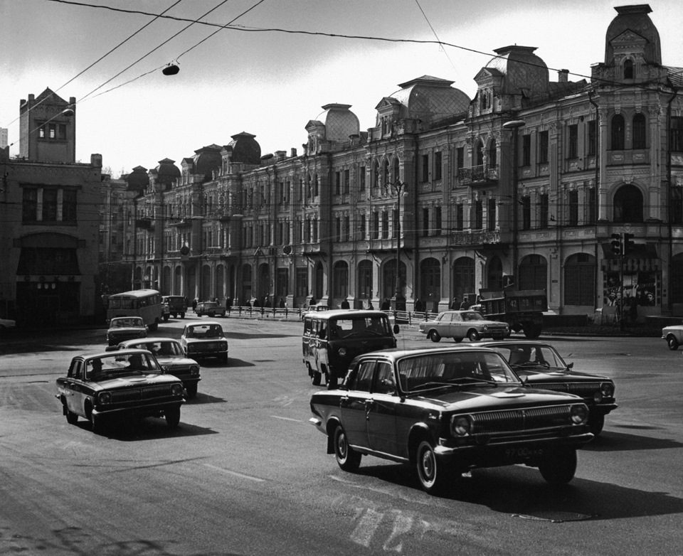 Historická ulica s autami v prevádzke a elegantnou architektúrou budovy, čiernobiela fotografia.