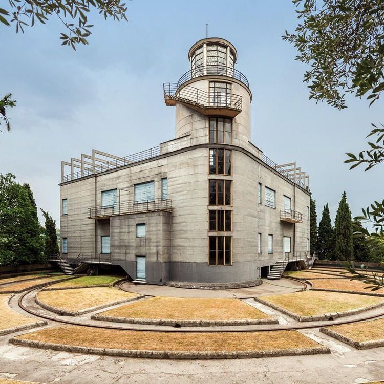 Moderná architektúra: betónová budova s vežou a panoramatickými oknami, zasadená do geometrickej záhrady.