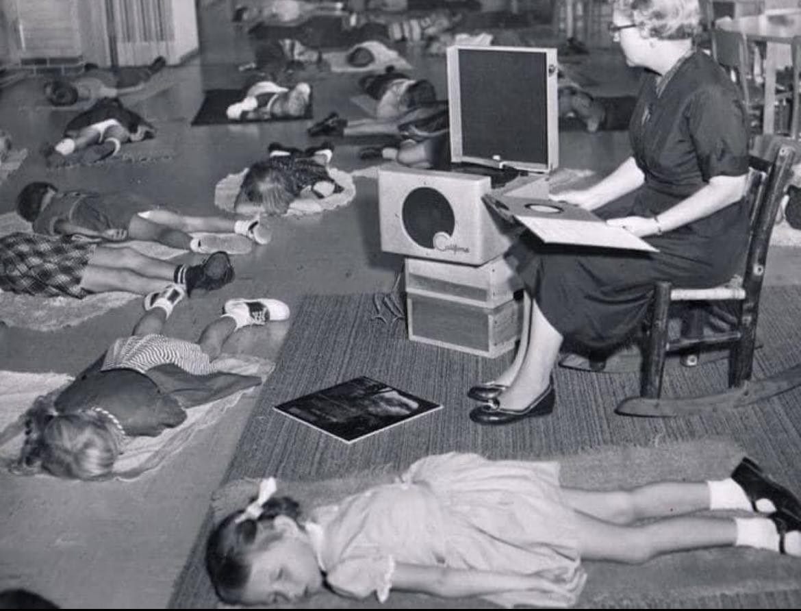 Deti ležia na podložkách v materskej škole, kde sa učiteľka stará o gramofón. Retro scéna z 50. rokov zachytáva odpočinok.