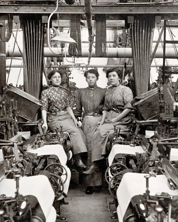 Tri ženy v historickej textilnej továrni medzi strojmi, čiernobiela fotografia, priemyselná revolúcia.