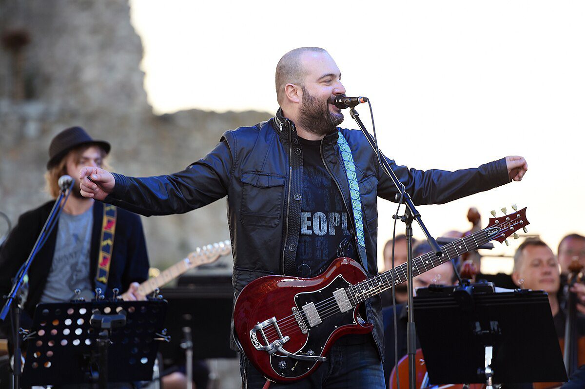 Hudobník s gitarou spieva do mikrofónu na vonkajšom koncerte. Publikum sleduje vystúpenie.