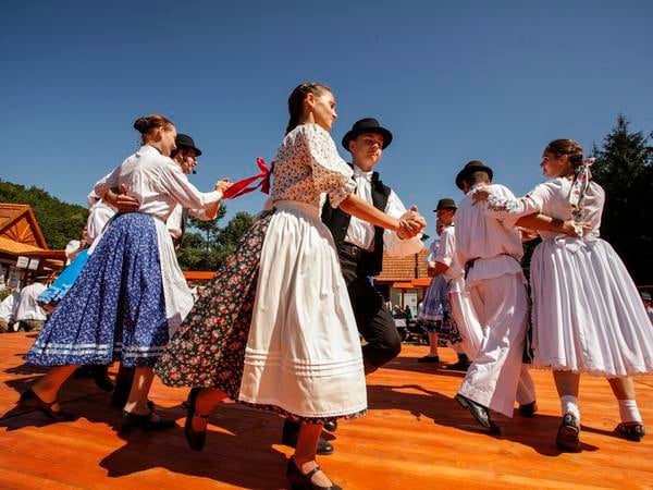 Skupina tanečníkov v krojoch tancuje na vonkajšej drevenej scéne pod modrou oblohou.