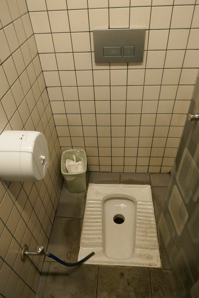 Verejné WC na štýl tureckého záchodu s kachličkovou stenou, košom, trubkou na vodu a nádržkou na splachovanie.
