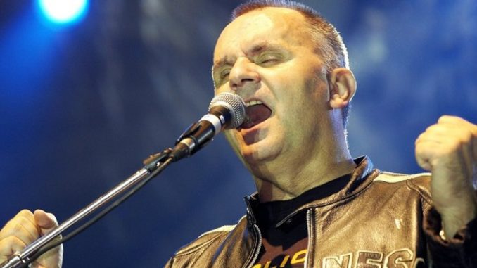 Ospravedlňujem sa, ale neviem, kto je na obrázku. Tu je alternatívny text: Spevák vystupujúci na pódiu s mikrofónom v koženej bunde.