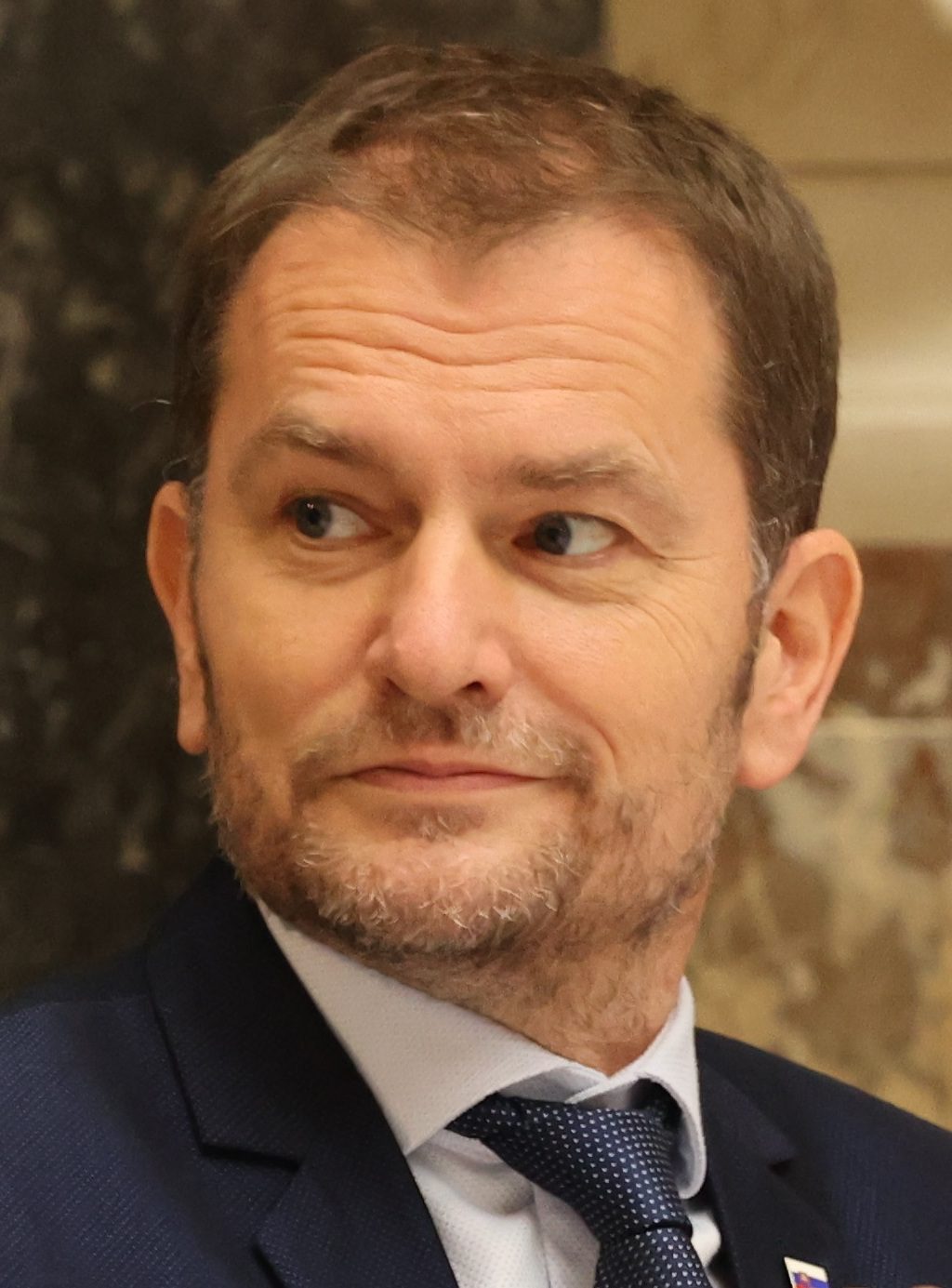 Loďujem, ale neviem, kto je na obrázku. Alternatívny text: Muž v modrom obleku a kravate pohľadom vľavo, interiér.