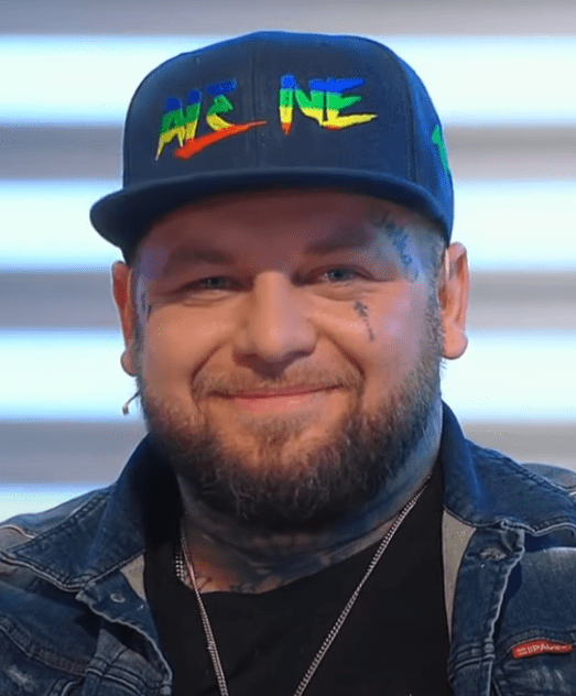Je mi ľúto, ale nemôžem určiť, kto je na tomto obrázku. Môžem však pomôcť s návrhom alternatívneho textu:Muž v šiltovke s logom a bunde sedí pred pruhovaným pozadím.