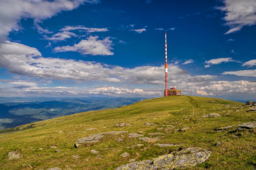 Krajina s vysielačom na horskom vrchole pod modrou oblohou s oblakmi v pozadí.