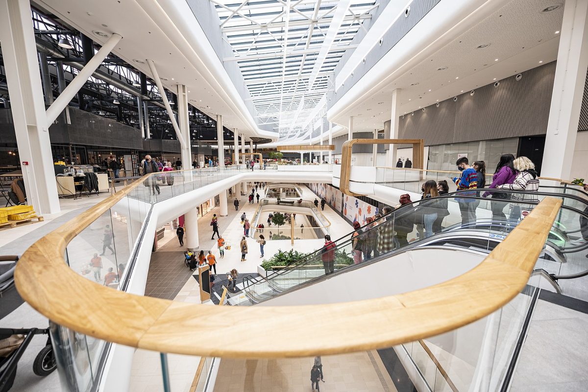 Moderné nákupné centrum s presklenou strechou a návštevníkmi na eskalátorech a poschodiach.