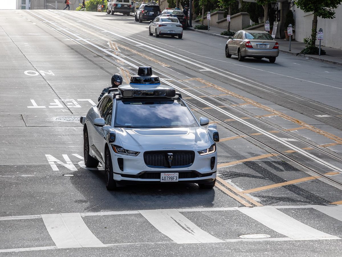 Biele autonómne auto na svahu v meste, vybavené LIDAR technológiou na streche, jazdí po prázdnej ceste.