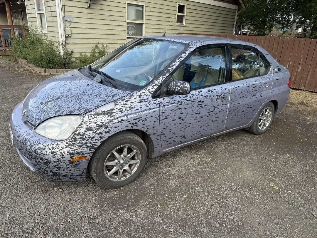 Šedé auto so špinavým blatovým vzorom na karosérii, zaparkované na prašnej ceste pri drevenom plote.