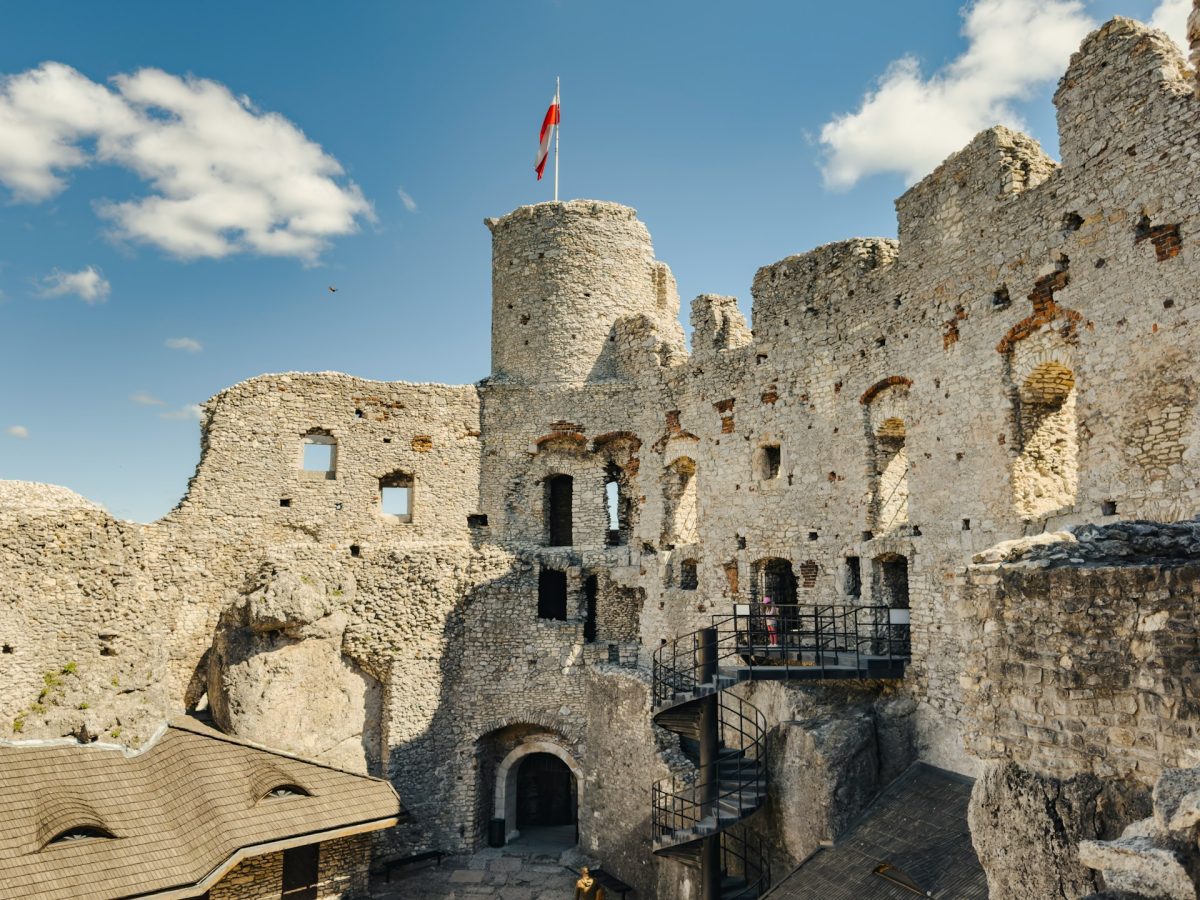 Stredoveký hrad so zástavou na veži, kamenné múry a schodisko pod modrou oblohou s oblakmi.