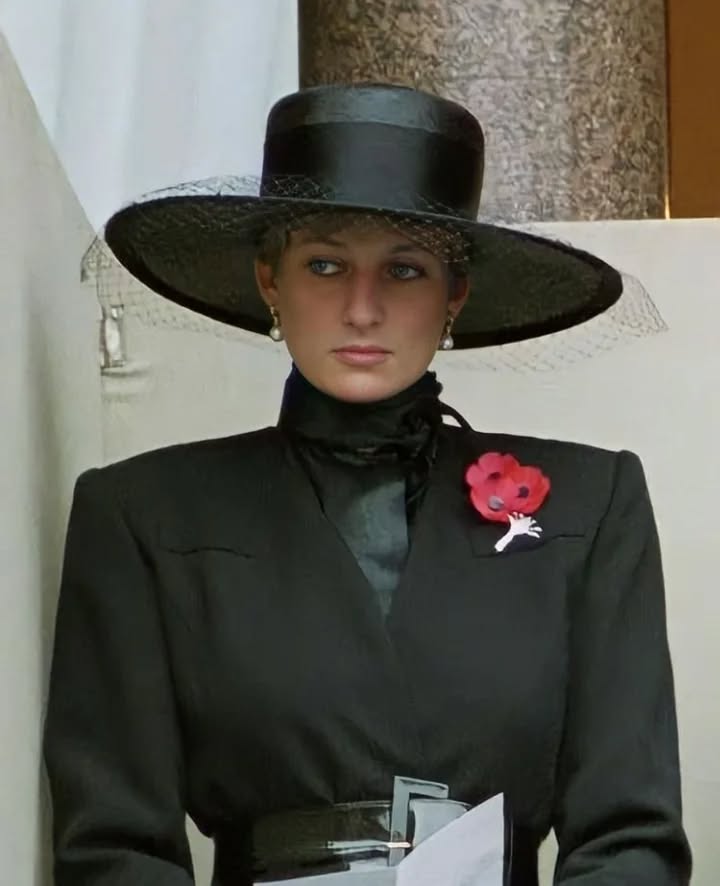 Osoba v elegantnom čiernom klobúku s červeným odznakom maku stojí s vážnym výrazom tváre.