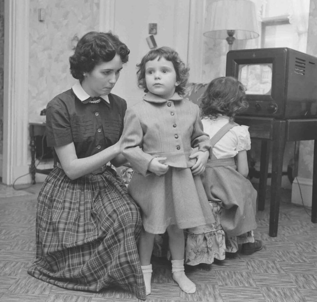 Matka so starostlivým pohľadom pomáha obliecť malú dcéru, zatiaľ čo iné dieťa sleduje televíziu v retro miestnosti.