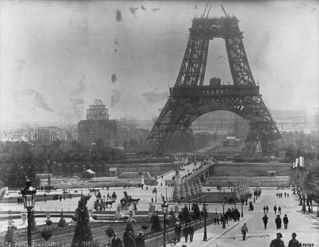 Historická fotografia Eiffelovej veže vo výstavbe, obklopená ľuďmi a parkom, Paríž, 19. storočie.