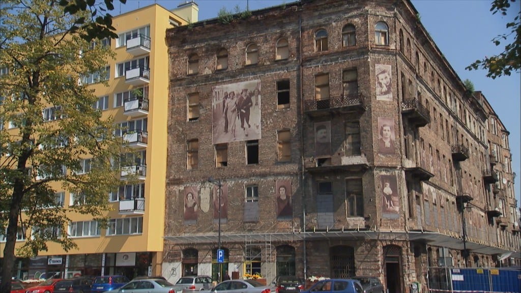 Historická tehlová budova s maľbami tvárí, vedľa moderného žltého domu, autá zaparkované pred budovami.