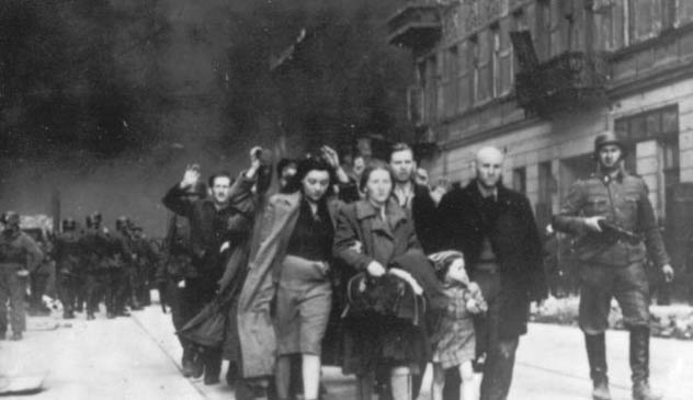 Skupina ľudí vedená vojakmi na ulici počas historickej udalosti, zachytená na čiernobielej fotografii.
