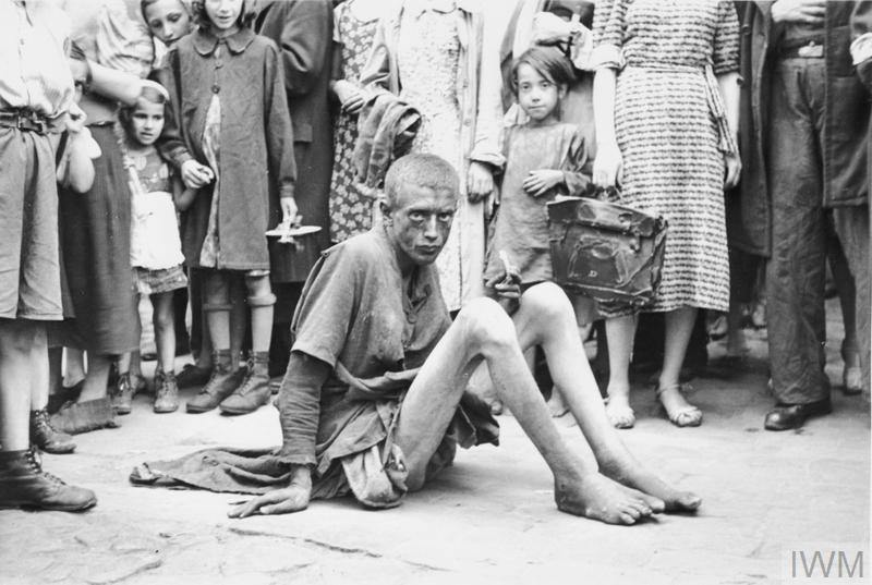 Ľudia v roztrhanom oblečení sedí na ulici obklopený davom, historické čiernobiele foto.