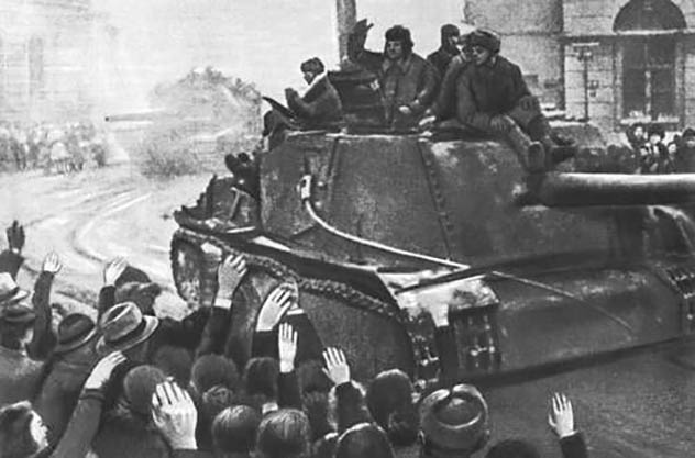 Skupina ľudí zdraví prechádzajúci tank na historickej ulici počas vojenského prechodu.
