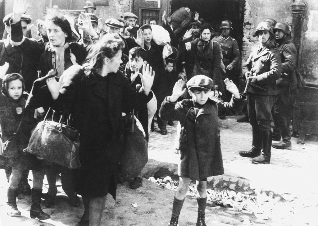 Ľudia so zdvihnutými rukami v sprievode vojakov v historickom kontexte druhej svetovej vojny.