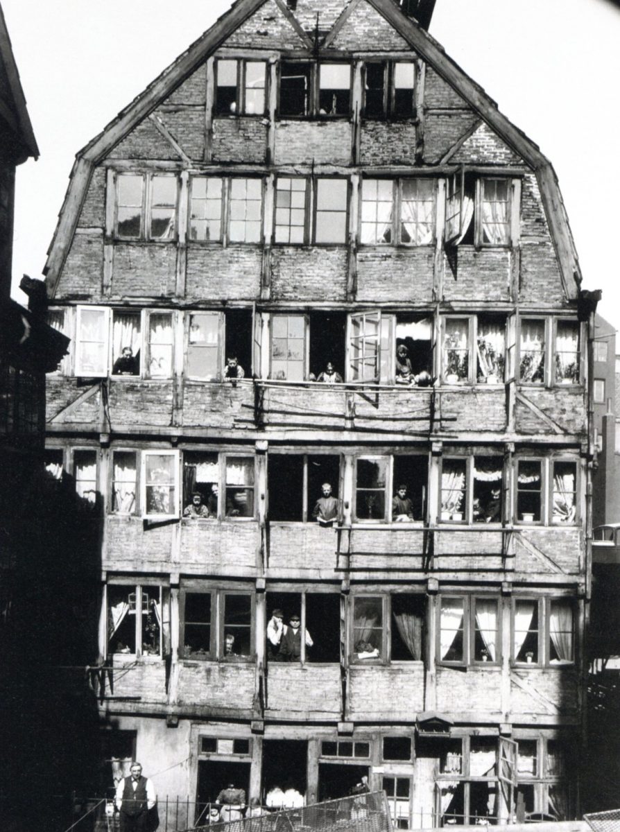 Starý viacposchodový drevený dom plný ľudí pozerajúcich z okien, historická architektúra mestského bývania.