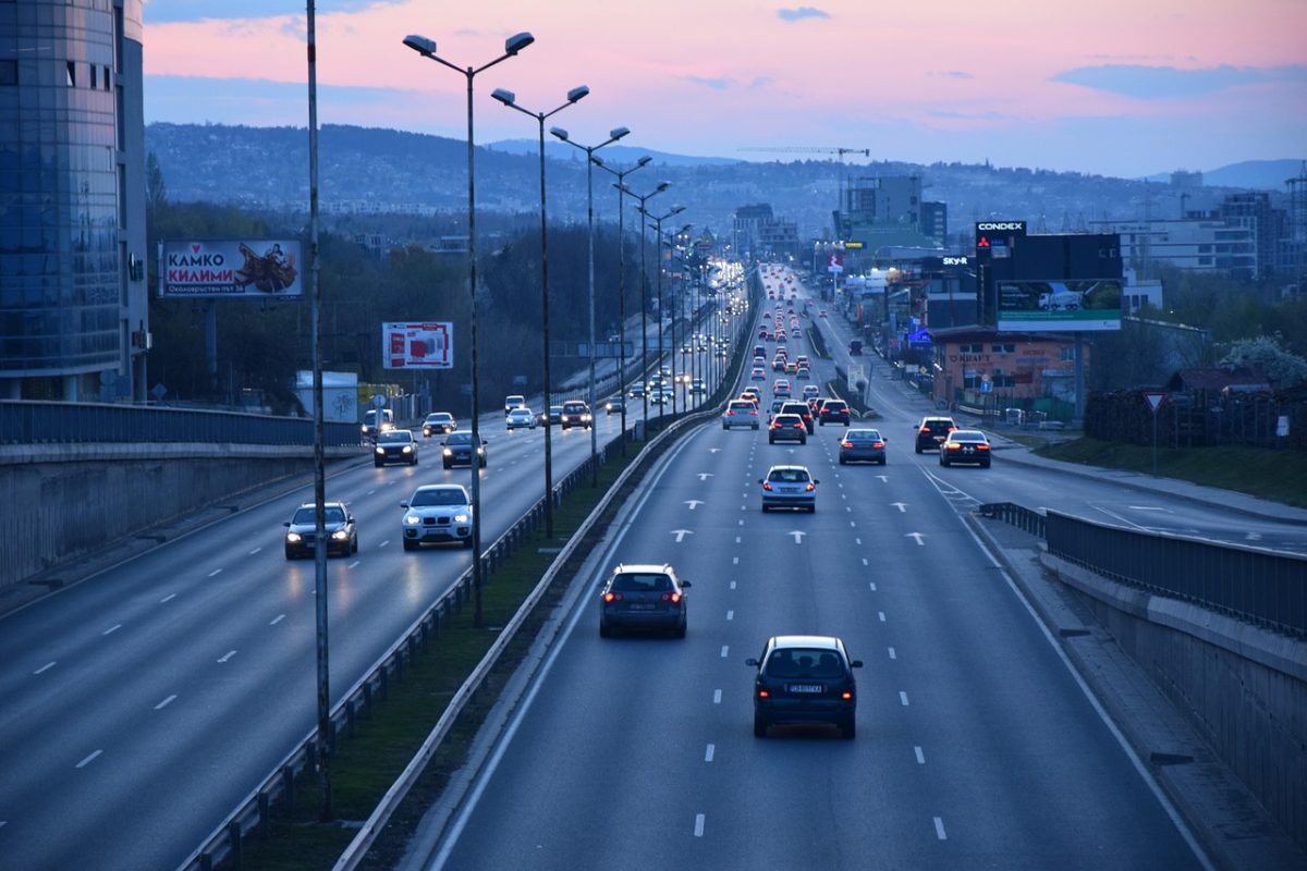 Večerná premávka na štvorprúdovej diaľnici s osvetlením a billboardmi, pohľad na mesto v diaľke.