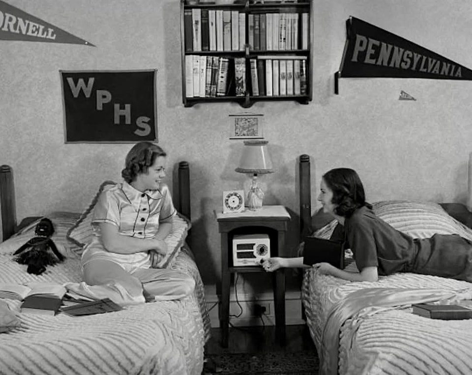 Dve ženy na internáte pri rozhovore v izbe, ozdobenej univerzitnými vlajkami, čiernobiela fotografia.
